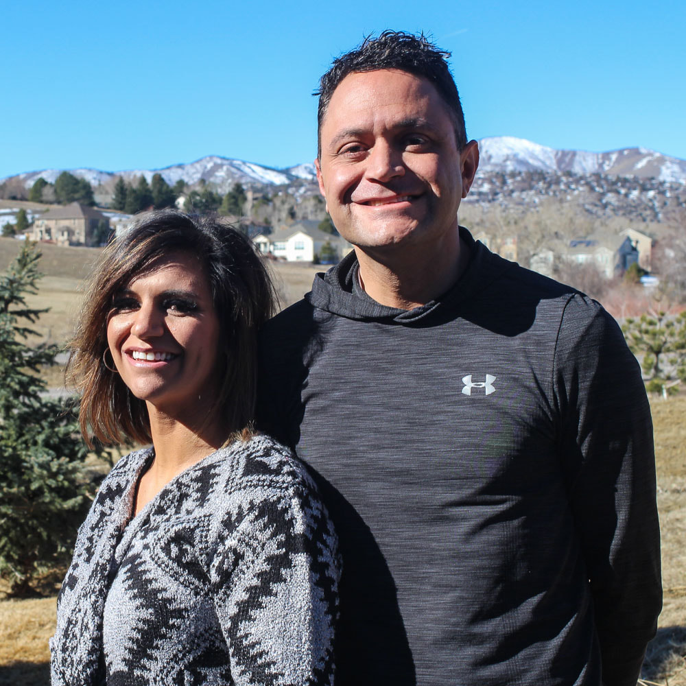 A Latinx brother and sister smile at the camera. They're standing on a hill, and behind them are the Rocky Mountains lightly dusted with snow.