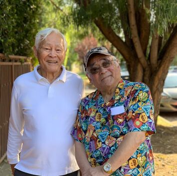 Two Asian American elderly men stand on a tree-lined street in Los Angeles. The man on the left wears a white shirt, which matches his white hair. The man on the right wears a baseball cap, sunglasses, and a multicolored polo shirt.