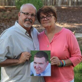 A middle-aged white couple stands in their backyard holding a large photo of their son, Alex. The man, left, wears a white and green collared shirt. The woman, right, wears a pink shirt. Both wear glasses.