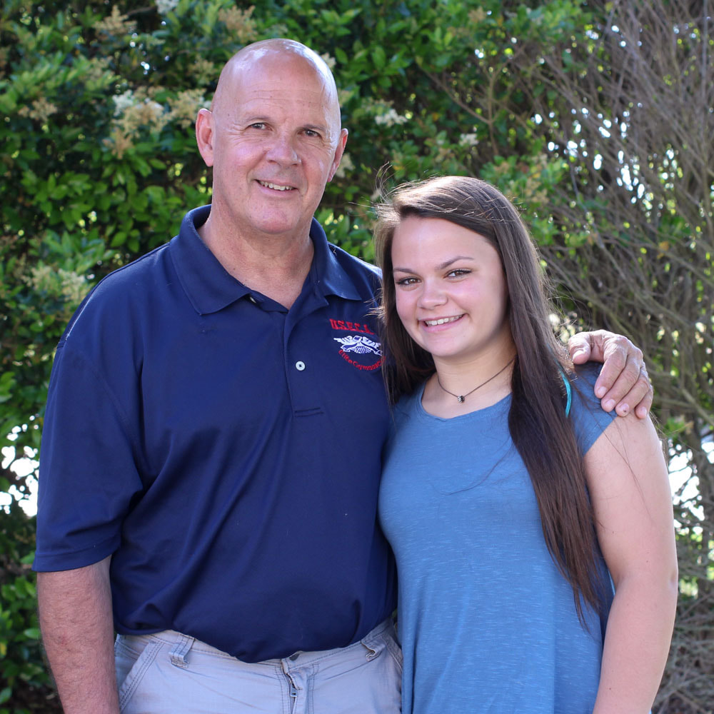 A white father and daughter smile at the camera, both wearing blue shirts. Behind them is green foliage.