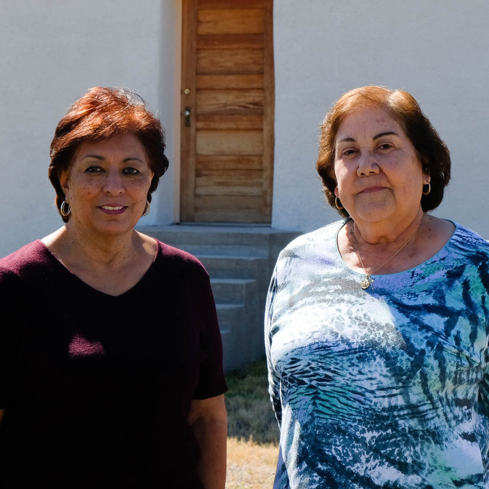 Two middle-aged Latina friends stand in front of a white adobe building on a sunny day.