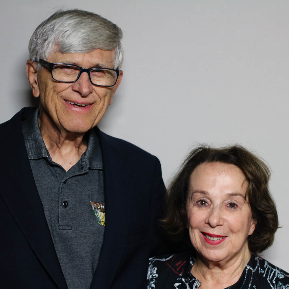 An elderly white couple smiles in front of a gray background. The man has black horn-rimmed glasses and is much taller than the woman, who wears red lipstick.