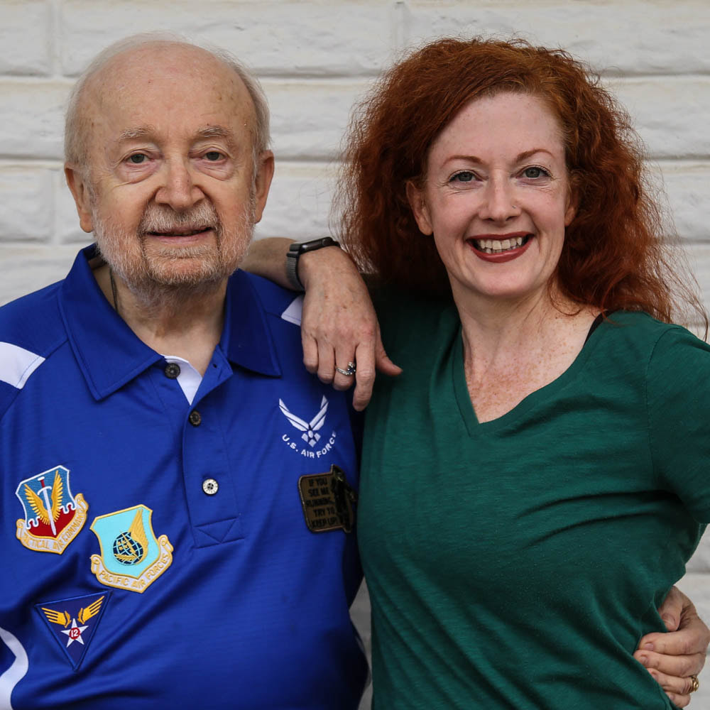 A white father and daughter smile in front of a white wall. The father is elderly and wears several military patches on his blue shirt. The daughter has bright red hair.