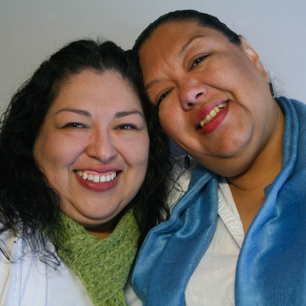 Two Latina sisters smile, their heads inclined toward one another. The sister on the left is wearing a green scarf. The sister on the right is wearing a blue silk scarf.